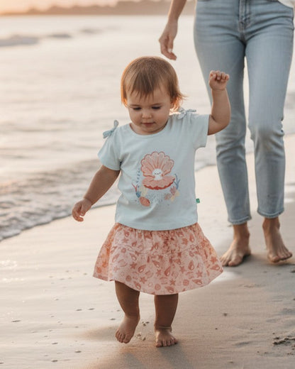 Toddler wearing a Calla Lily Cosmetics + CL kids PlayBud set with a seashell graphic top and patterned skirt on a beach