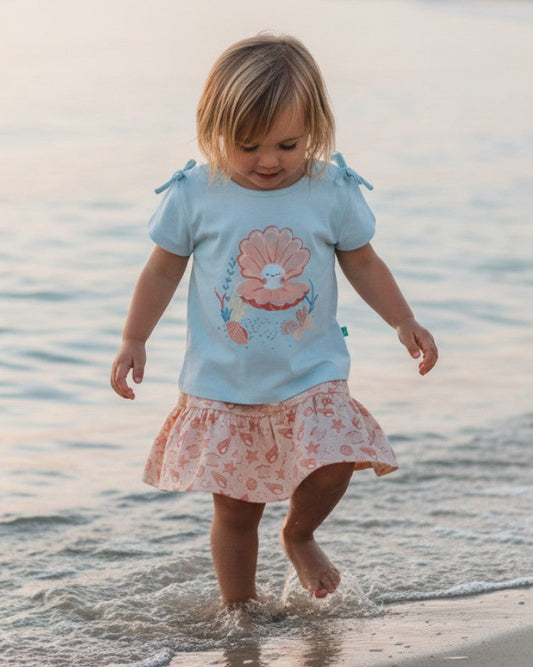 Toddler wearing a Calla Lily Cosmetics + CL kids PlayBud set with a blue shell-print top and matching skirt at beach