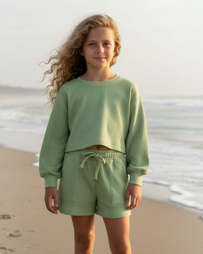 Tween girl wearing a sage waffle knit two-piece set from Calla Lily Cosmetics + CL kids on a beach at sunset.