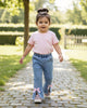 Toddler wearing Calla Lily Cosmetics + CL kids blue jeans with raw hem, pearl accents, and pink bows on a path.