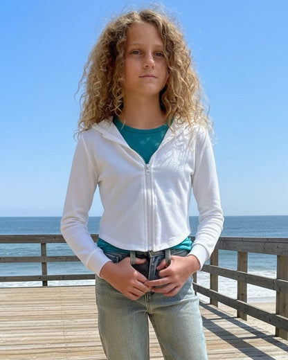 Girl wearing a white French Terry Crop Zip Up Hoodie by Calla Lily Cosmetics + CL kids on a wooden pier by the ocean.