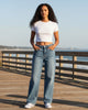 Model wearing Calla Lily Cosmetics + CL kids YMI low rise wide leg blue denim jeans with a white crop top on a pier.