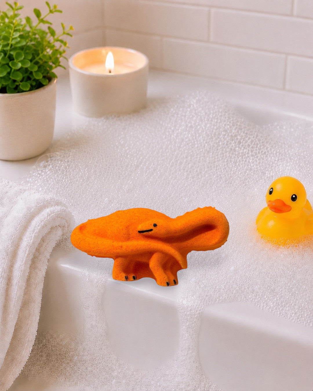 Calla Lily Cosmetics + CL kids orange brontosaurus bath bomb on a tray with spa decor, towels, and a lit candle.