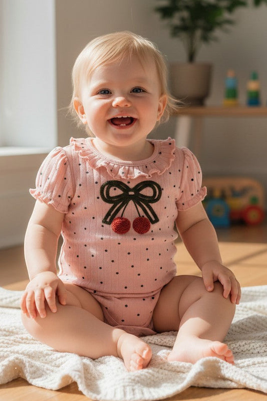 Calla Lily Cosmetics + CL kids pink cotton bodysuit with black polka dots, ruffled collar, and cherry bow applique. Lap shoulders and snap closure.