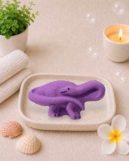 Calla Lily Cosmetics + CL kids purple brontosaurus dinosaur bath bomb with grape scent on a white wood background.