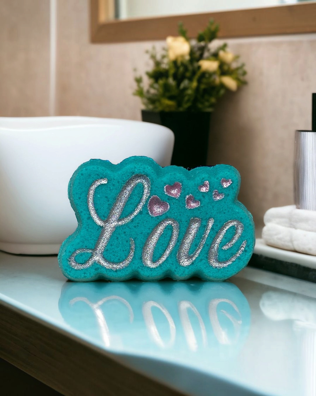 Calla Lily Cosmetics + CL kids teal Love bath bomb with silver glitter lettering and pink heart accents on a vanity.