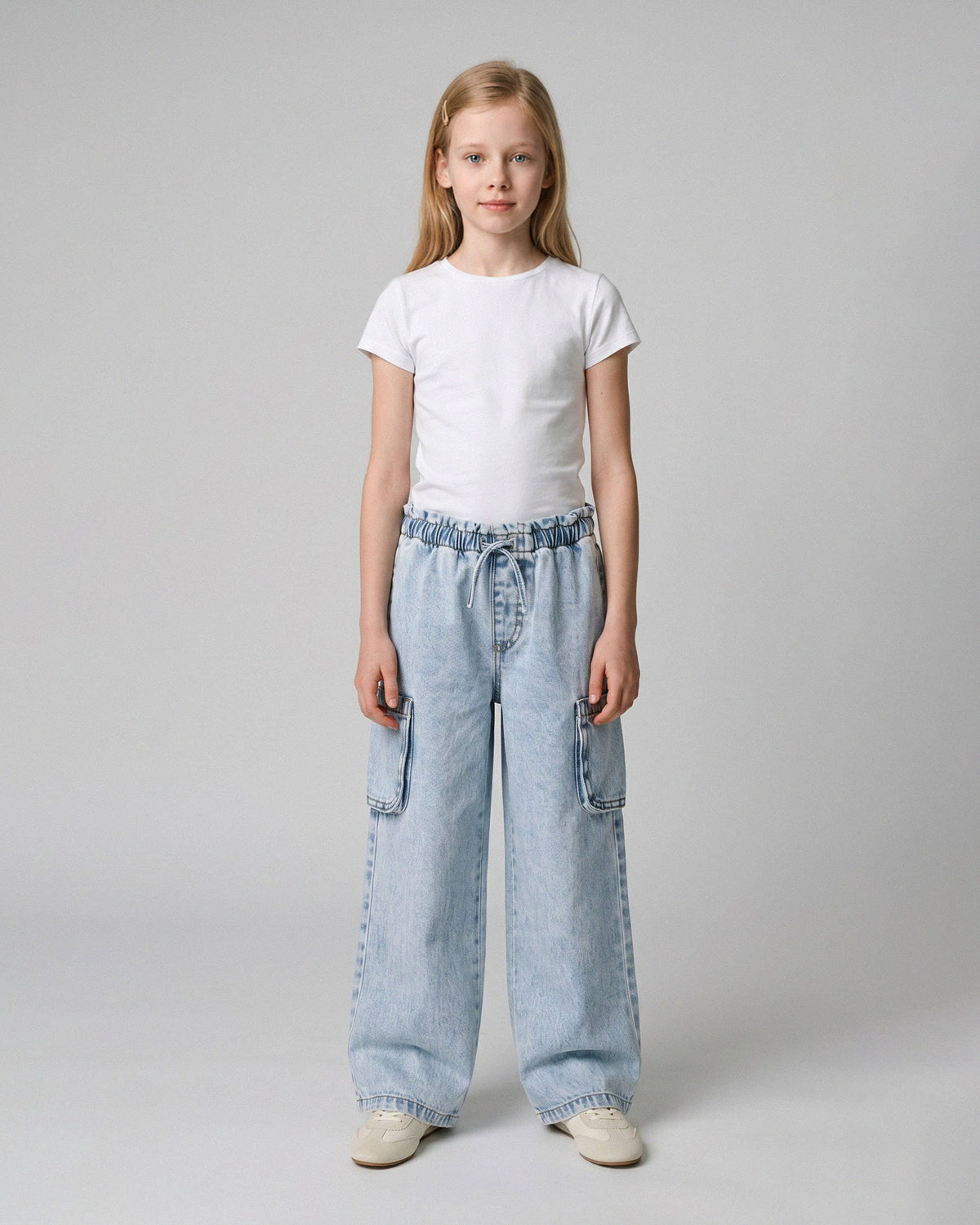 A young girl stands against a light gray studio background wearing a plain white short-sleeve T-shirt, light-wash wide-leg denim cargo pants, and off-white sneakers.