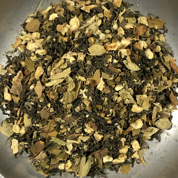 A close-up top-down view of loose-leaf herbal tea or spice blend in a metal bowl, showing dark dried leaves mixed with light tan and beige botanical pieces.