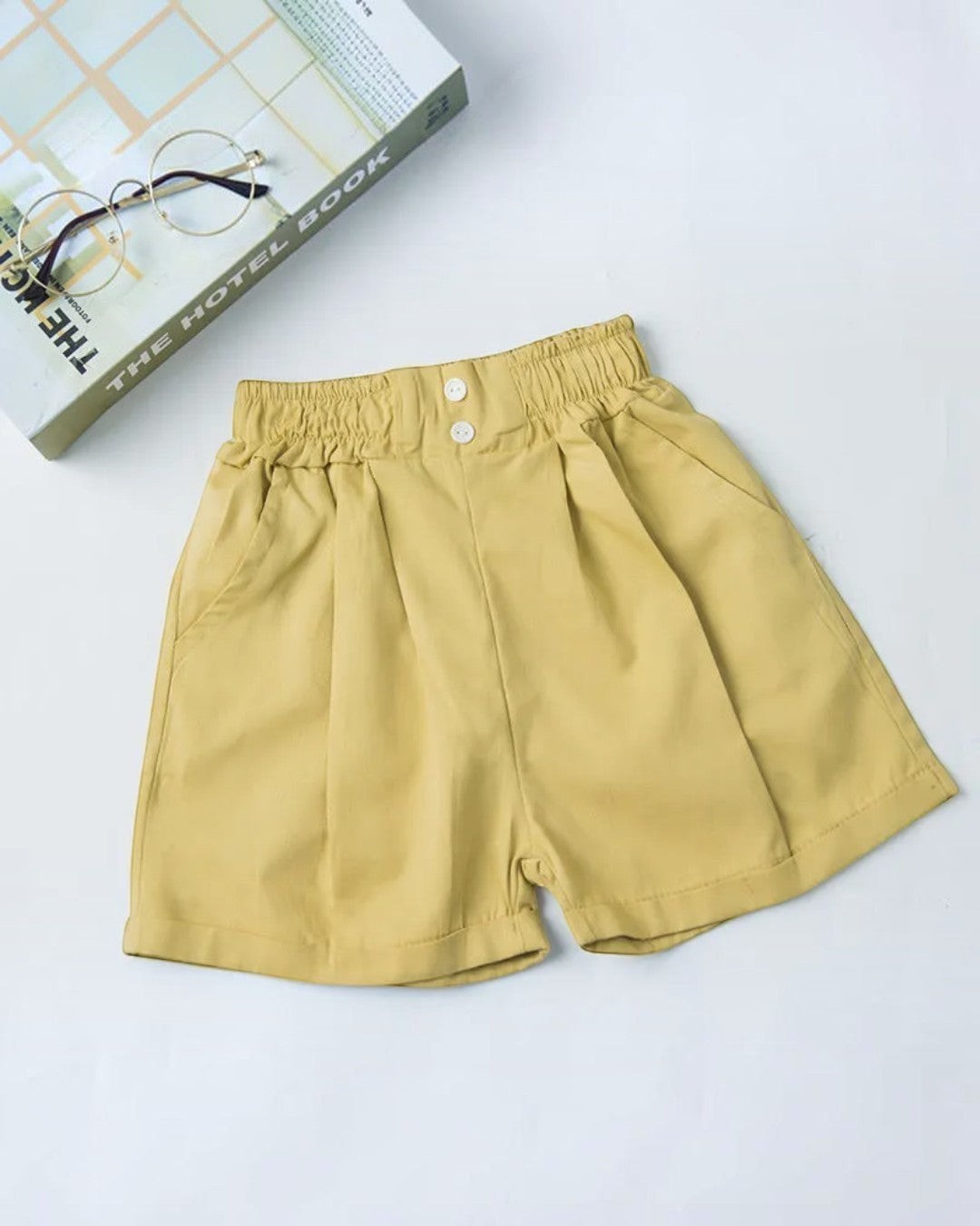 Beige shorts on a light gray background with a book and glasses in the corner.
