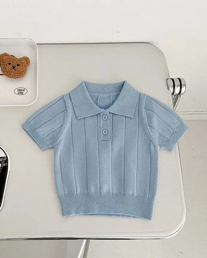 Light blue children's shirt on a white surface with black shoes and a teddy bear in the background.