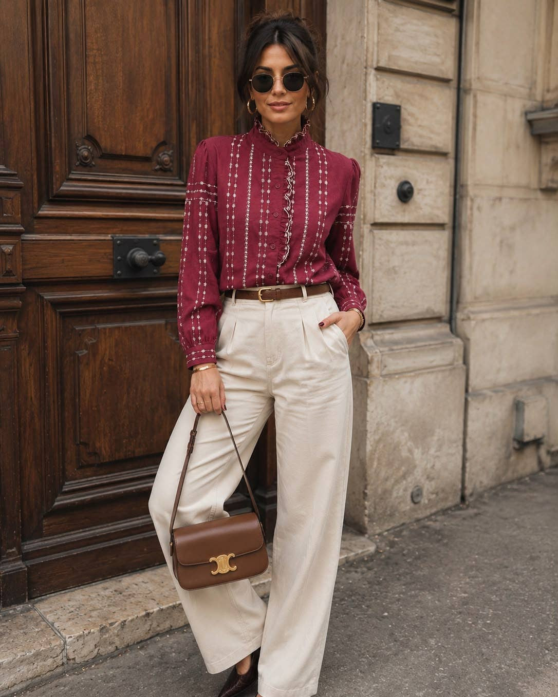 A woman stands on a city street wearing a burgundy long-sleeve button-up shirt with white vertical embroidery, cream wide-leg trousers, a thin brown belt, dark pointed flats, and a brown leather crossbody bag.
