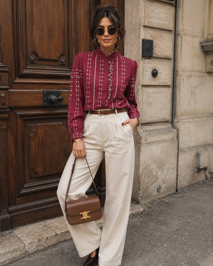 A woman stands on a city street wearing a burgundy long-sleeve button-up shirt with white vertical embroidery, cream wide-leg trousers, a thin brown belt, dark pointed flats, and a brown leather crossbody bag.