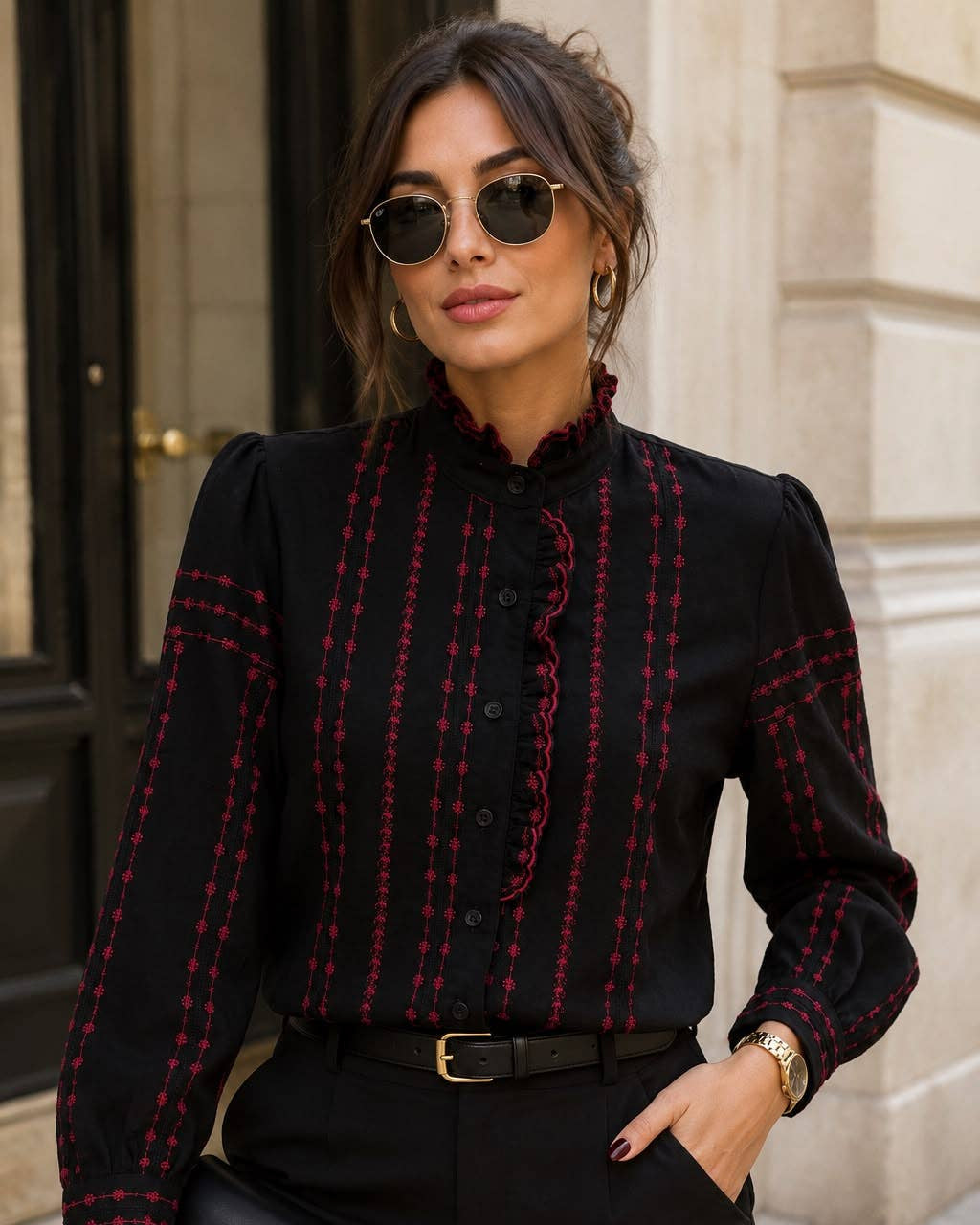 A woman wearing a black long-sleeve blouse with red vertical stripe patterns and ruffle details, paired with black trousers and holding a black handbag, set against a stone building background.