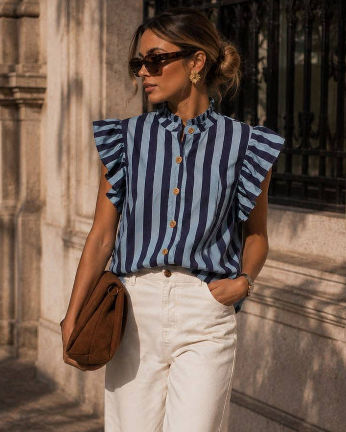 A woman stands on a sunlit stone sidewalk wearing a blue and white vertically striped ruffle-sleeve blouse, white wide-leg trousers, brown shoes, and carrying a brown shoulder bag.