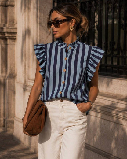 A woman stands on a sunlit stone sidewalk wearing a blue and white vertically striped ruffle-sleeve blouse, white wide-leg trousers, brown shoes, and carrying a brown shoulder bag.