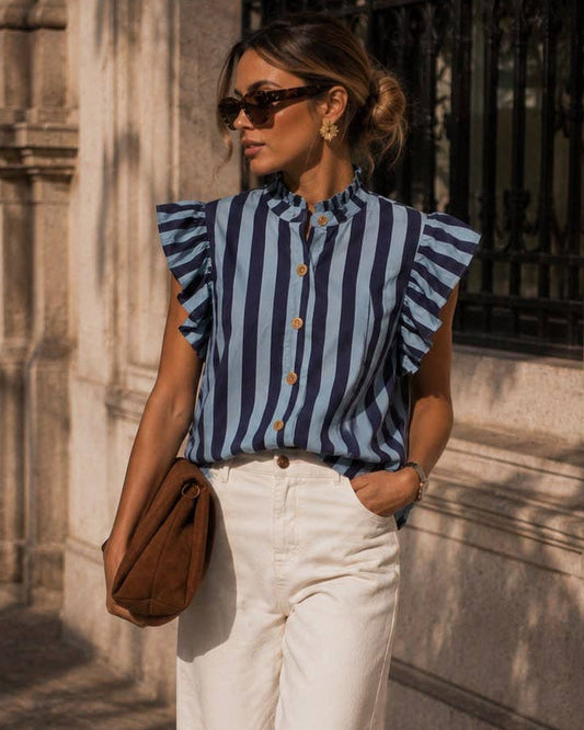A woman stands on a sunlit stone sidewalk wearing a blue and white vertically striped ruffle-sleeve blouse, white wide-leg trousers, brown shoes, and carrying a brown shoulder bag.