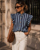 A woman stands on a sunlit stone sidewalk wearing a blue and white vertically striped ruffle-sleeve blouse, white wide-leg trousers, brown shoes, and carrying a brown shoulder bag.