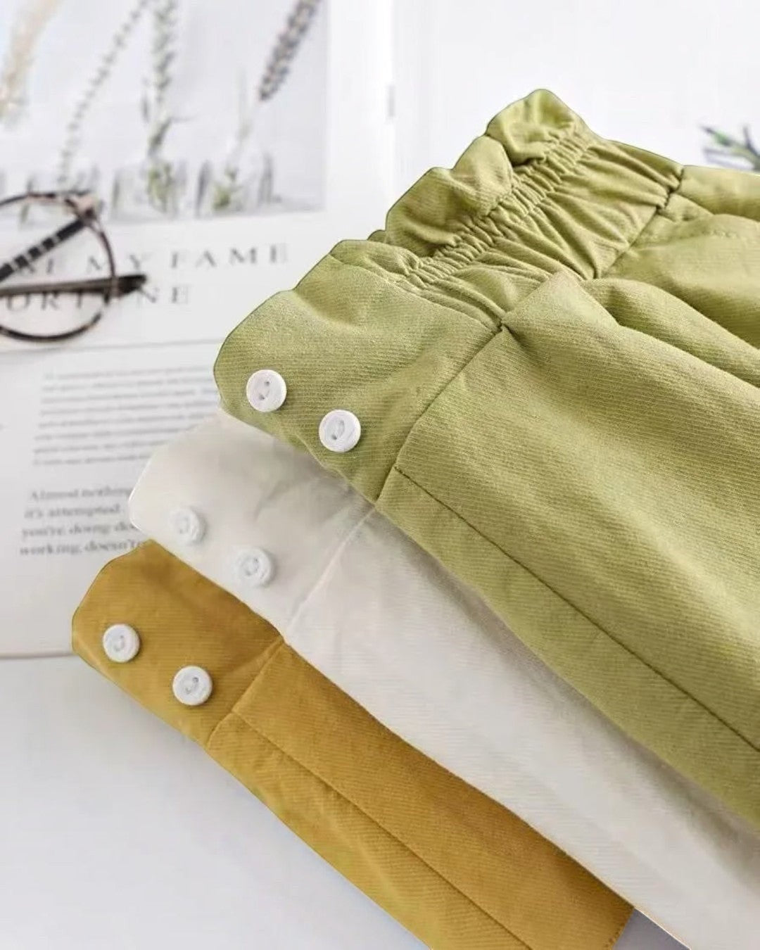 Folded green and mustard yellow pants with white buttons on a light surface.