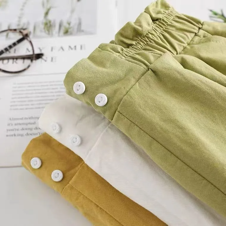Folded green and mustard yellow pants with white buttons on a light surface.