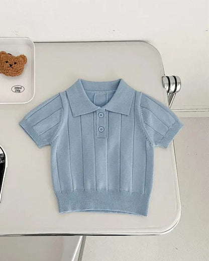 Light blue children's shirt on a white surface with black shoes and a teddy bear in the background.