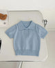 Light blue children's shirt on a white surface with black shoes and a teddy bear in the background.