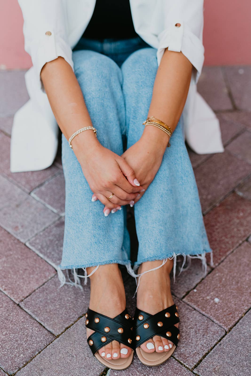 Person wearing black sandals with gold studs, blue jeans, and a white jacket on a brick pavement.