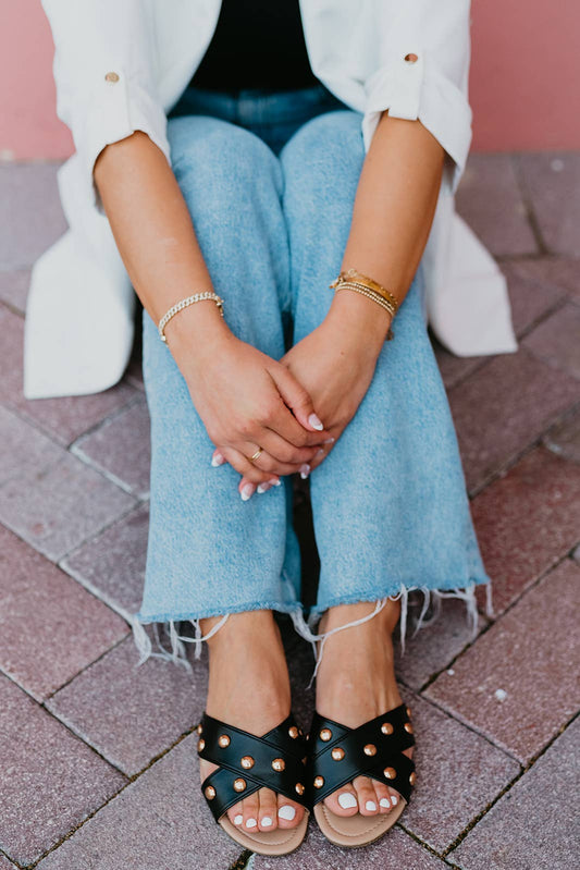 Person wearing black sandals with gold studs, blue jeans, and a white jacket on a brick pavement.