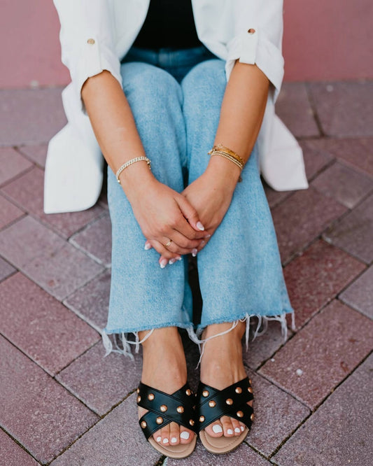 Person wearing black sandals with gold studs, blue jeans, and a white jacket on a brick pavement.