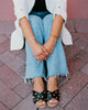 Person wearing black sandals with gold studs, blue jeans, and a white jacket on a brick pavement.