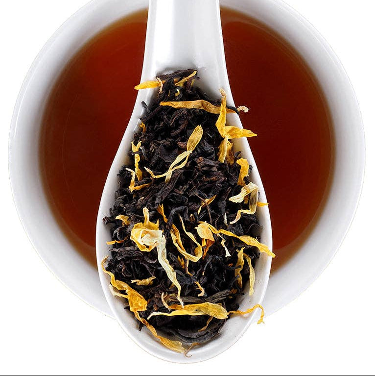 A white ceramic spoon resting in a white cup filled with amber-brown tea, with loose dried tea leaves and yellow flower petals visible in the spoon. The Find Elmwood Tea Inn logo appears in the top right corner.