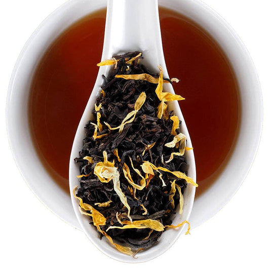 A white ceramic spoon resting in a white cup filled with amber-brown tea, with loose dried tea leaves and yellow flower petals visible in the spoon. The Find Elmwood Tea Inn logo appears in the top right corner.