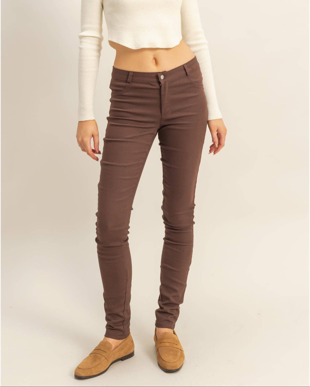 A woman modeling slim-fit brown skinny jeans with a cream ribbed long-sleeve crop top and tan suede loafers, standing against a plain beige studio background.
