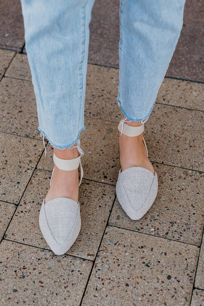 White pointed-toe shoes worn with light blue jeans on a tiled floor.