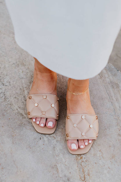 Beige sandals with pearl embellishments worn by a person on a light stone surface.