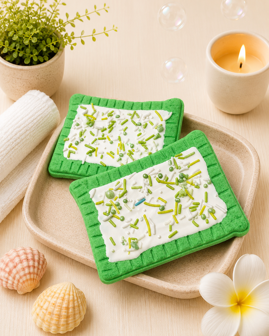Two green coasters with a white and green pattern on a wooden surface with a candle and plant in the background.