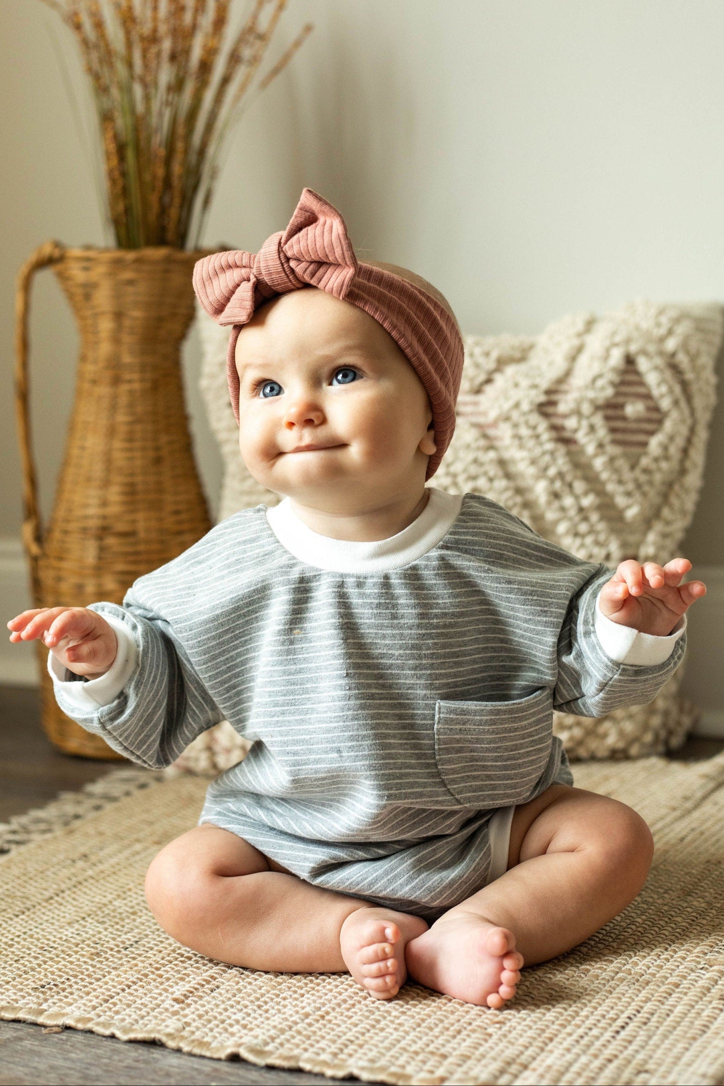 Baby sitting on a rug wearing a striped outfit and pink headband, with a woven basket and decorative items in the background.