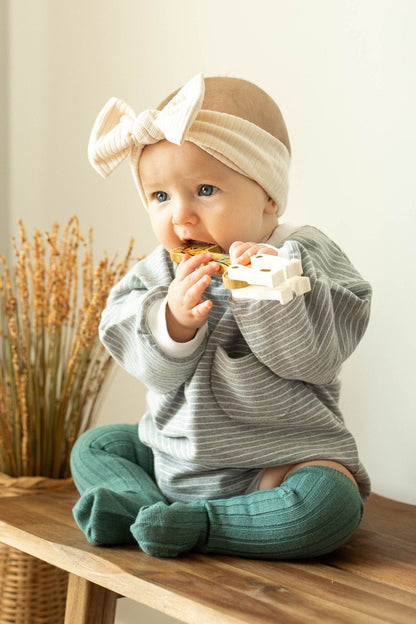 Baby sitting on a wooden surface wearing a gray sweater and green pants, with a beige headband.