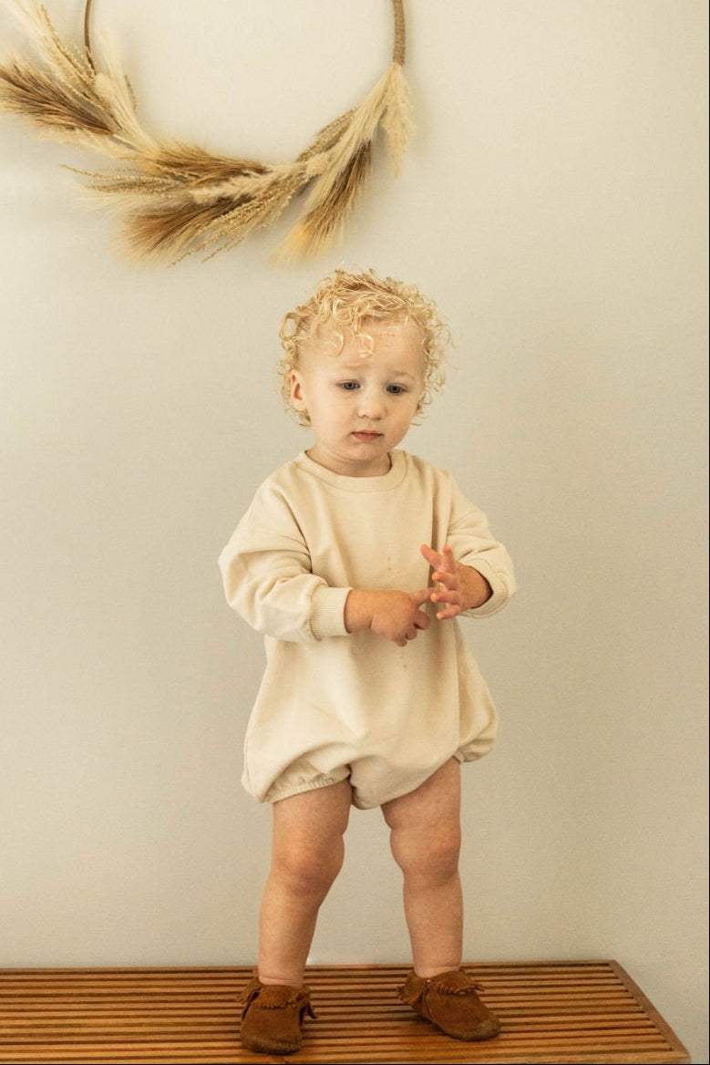 Child standing on a wooden platform with a decorative wreath of dried grasses on a plain wall.