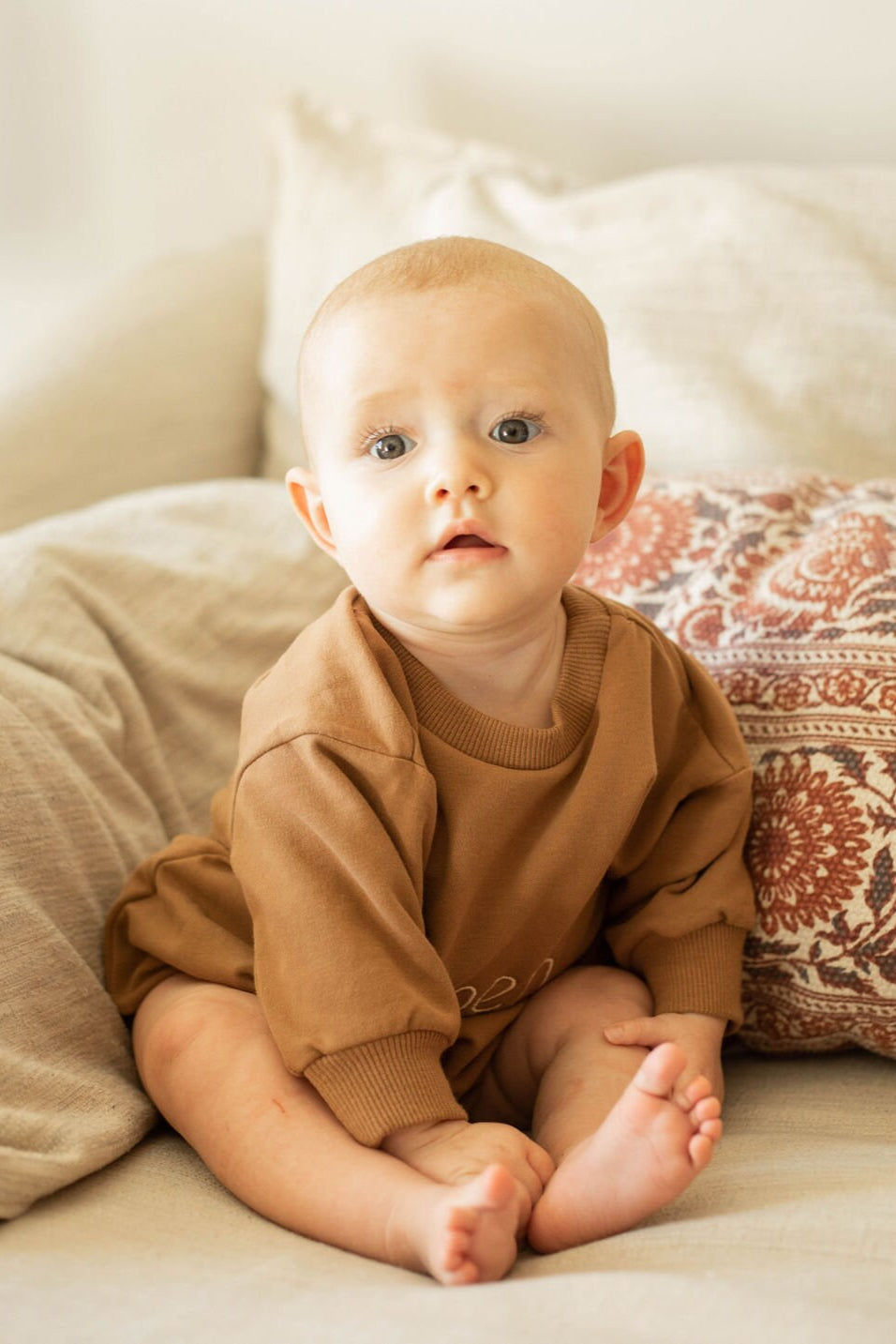 Baby in a brown outfit sitting on a couch with a patterned pillow.