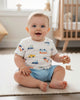 Baby sitting on a rug in a nursery with toys and a crib in the background