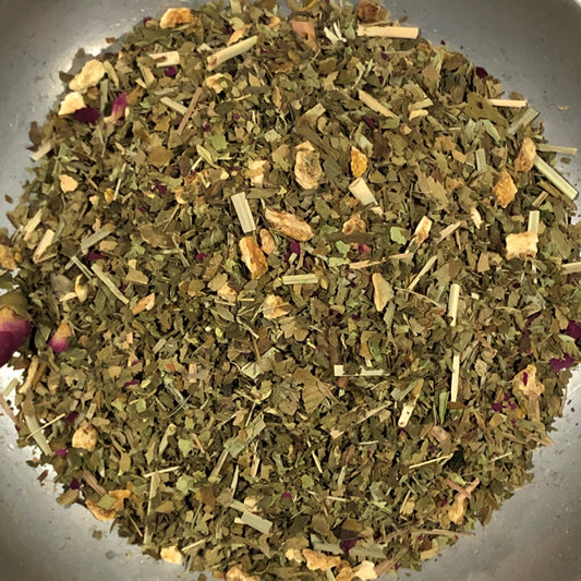 A close-up top-down view of loose-leaf herbal tea mixed with dried botanicals, including green leaves, light tan sticks, and small purple flower pieces, piled in a clear container.