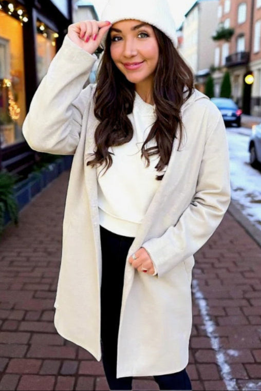 Woman wearing a long cream coat and white beanie on a snowy street.