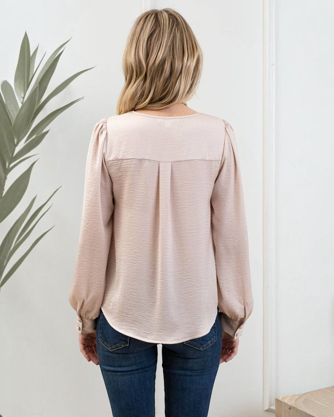 Person wearing a beige blouse and blue jeans standing against a white wall with a plant to the left.