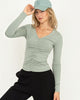 Woman wearing a green long-sleeve top and black pants with a light gray cap on a white background