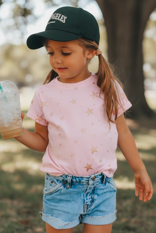Child in a park wearing a pink shirt, denim shorts, and a black cap holding a Starbucks drink.