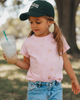 Child in a park wearing a pink shirt, denim shorts, and a black cap holding a Starbucks drink.