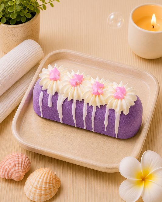A purple rectangular bath stone or soap bar decorated with white cream drips and pink flower accents, displayed on a beige tray with spa-themed props including a candle, plant, shells, and a flower.