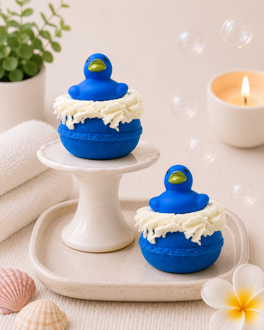 Two blue rubber duck-shaped bath bombs with white frosting on a white surface with a candle and plant in the background.