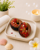 Two chocolate donuts with red and yellow toppings on a white plate, surrounded by bubbles, a candle, and decorative items.
