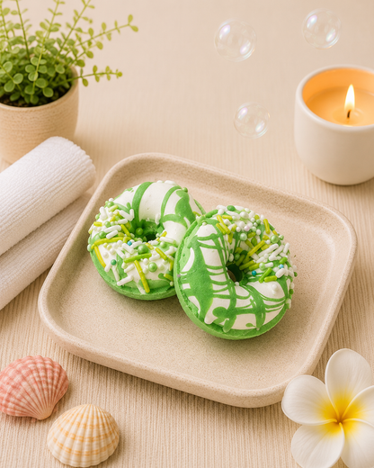 Two green and white donut-shaped soaps on a beige plate with a candle, shells, and a plant in the background.